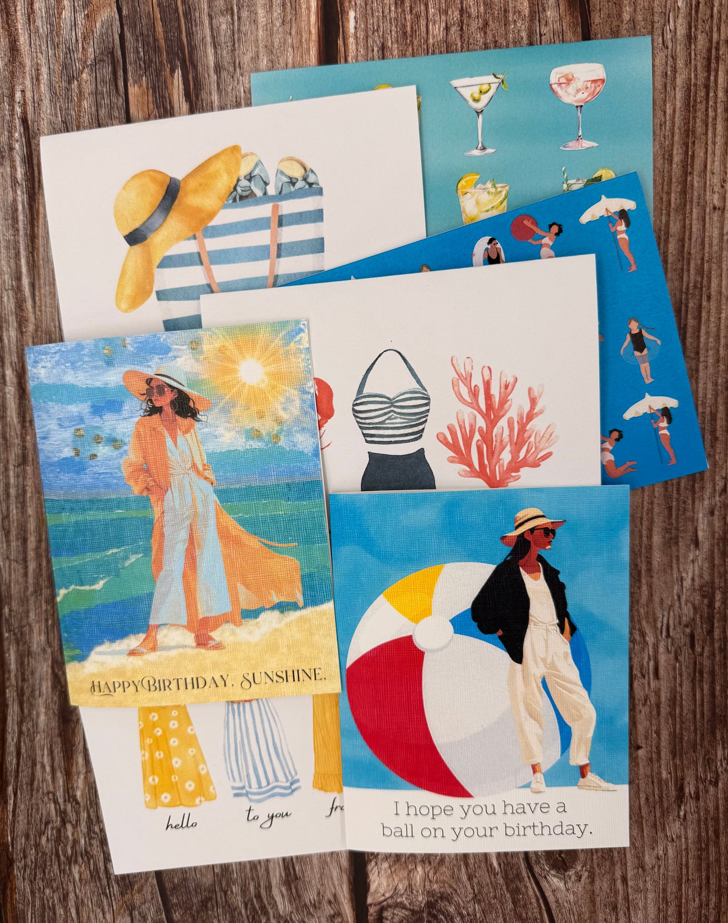 Seasonal Notecards by INNRETROspect displayed on a wooden table. These cards are playful and showcase imagery of summer. Featuring a giant beach ball, seascapes, and beach bags. 