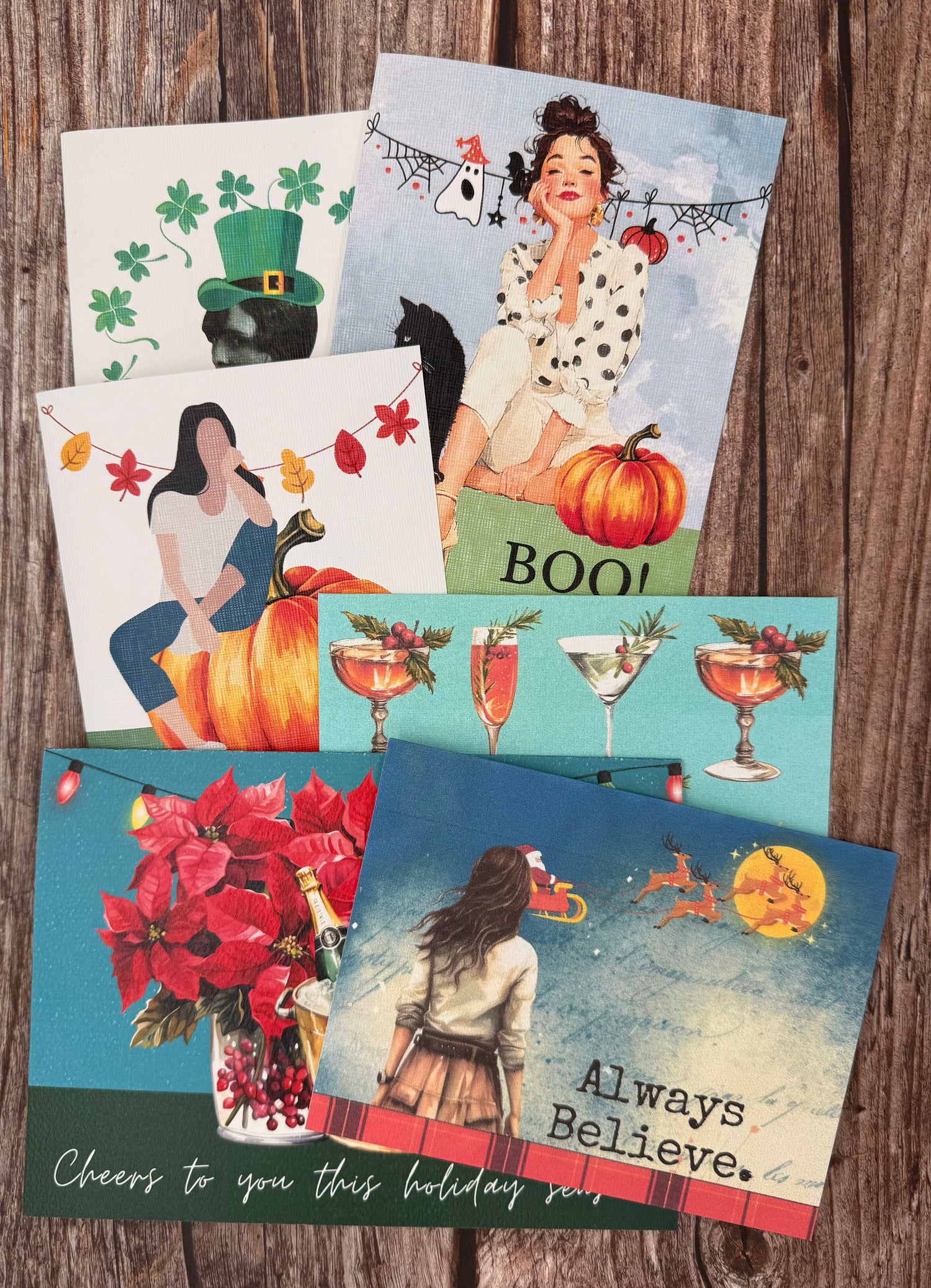 Flat lay of  cards that feature holiday cards for St.Patty's Day, Halloween, and Christmas. The bottom two cards read "cheers to you this holiday season" and "always believe" with a girl looking at Santa on his sleigh. 