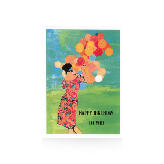 This card features an illustration of a lady in a bright printed dress from behind.  She has her hair up on her head with a blue scarf and is holding a huge bunch of multi-colored balloons.  The scene is an abstract grass and sky.  Happy Birthday to you is printed in all caps to the right of the lady on the grassy area
