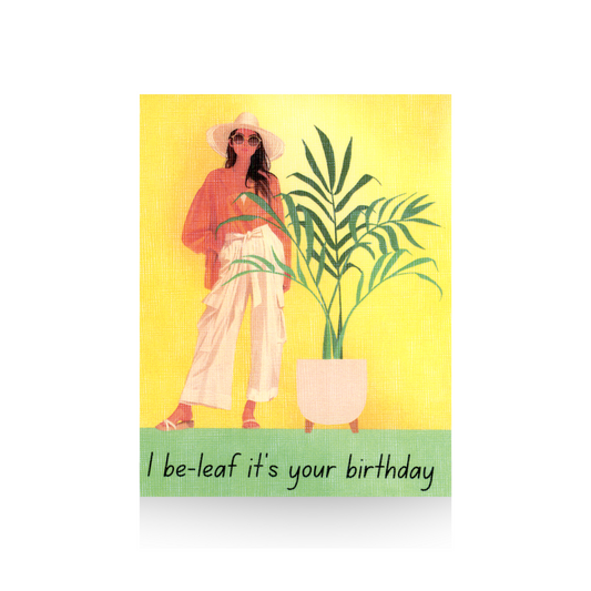 This birthday card features a modern illustrated lady in white wide-legged pants and brown long-sleeved top.  She is wearing sandals, sunglasses and a big hat.  To the right of her is a large palm plant in a white planter.  The background is yellow and the base is green.  The greeting "I be-leaf it's your birthday" is written on the base of the card