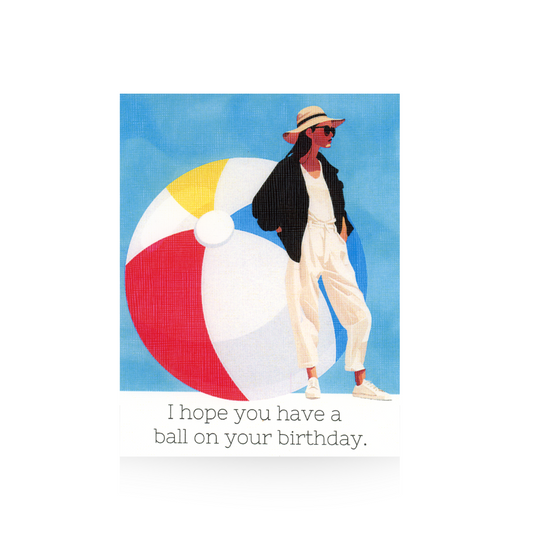 This birthday card features an oversized multi-colored beach ball against a blue sky on a white base.  Standing to the right of the beach ball is an illustrated modern lady in white pants and tennis shoes and b lack jacket.  She has grown hair and is wearing sunglasses and a wide-brimmed hat.  The message "I hope you have a ball on your birthday" is written on the bottom of the card