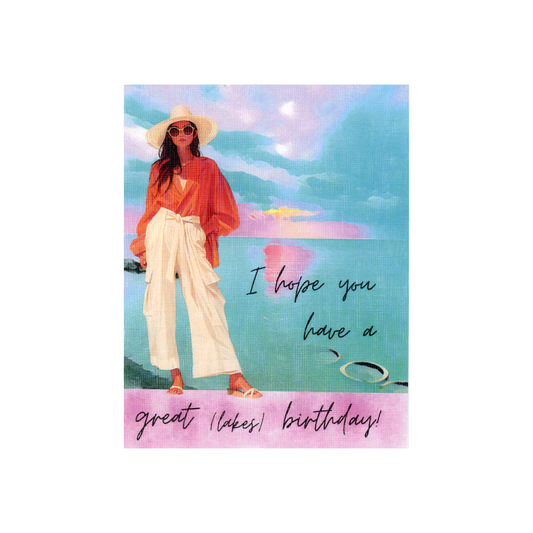 This birthday card features a watercolor-inspired scene of a Great Lake in colors of aqua, blue, and pink, with a modern lady standing by the lake.  She is wearing sandals, wide-legged cream pants, a rust-colored blouse, sunglasses and a hat.  She has long dark hair.  The message "I hope you have a great (lakes) birthday is written on the water and at the bottom of the card in cursive font.  The bottom banner is pink. 
