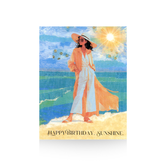 This birthday card features a hand-collaged background of the ocean and beach with a bright sun in top right hand corner.  A modern illustrated lady is standing on the beach in a white pantsuit and long tan jacket.  She has long brown hair, is wearing sunglasses and a wide-brimmed hat.  The message "Happy Birthday, Sunshine" is written on the bottom of the card