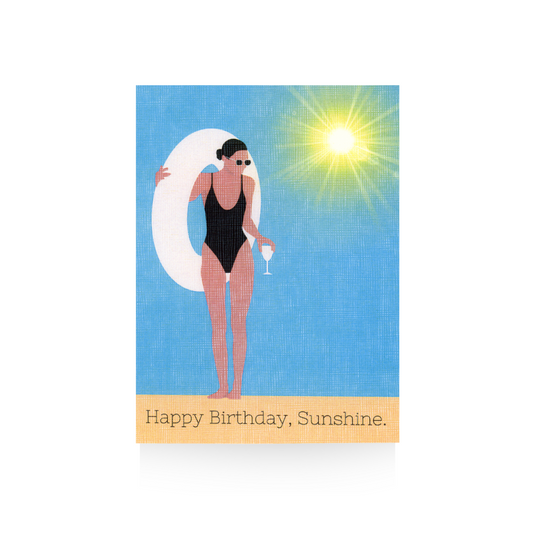 This birthday card features a modern illustrated lady in a black one-piece bathing suit holding a white inter tube in one hand and a cocktail in the other.  The background is sky blue and there is a bright sun in the top right hand corner.  The bottom is tan and features the greeting "Happy Birthday, Sunshine."