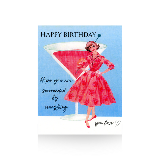 This birthday card features a blue background and white base upon which an oversized pink cocktail sits.  Standing to the right of the cocktail is a vintage lady in a pink print dress with a full skirt.  The greeting "HAPPY BIRTHDAY - Hope you are surrounded by everything you love" is written on the card.
