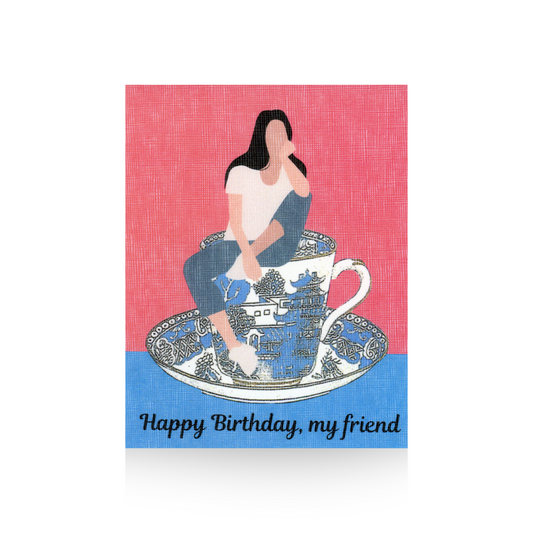 This friend birthday card features a vintage blue willow teacup on a blue base with a pink background.  Sitting on the teacup is a modern illustrated lady in blue jeans and a white t-shirt.  She has one leg propped up and her elbow is resting on her knee and her hand is resting on her chin.  The greeting "Happy Birthday, my friend" is written on the base of the card