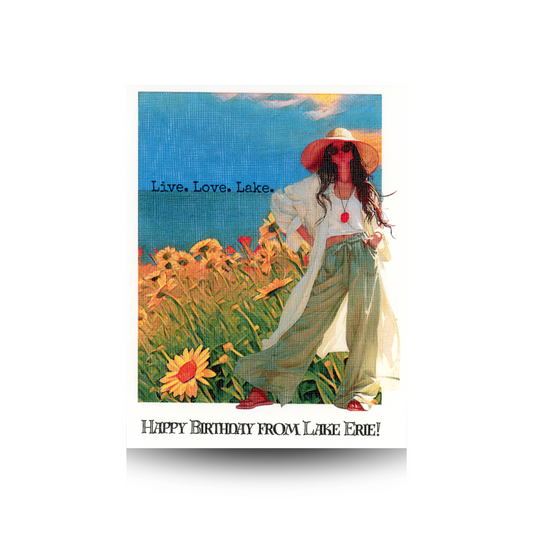 This birthday card features a modern woman with wide-legged pants, sunglasses, and a floppy hat with her hands on her hips, standing beside a hill of bright yellow flowers with the blue water of the lake and the blue sky behind her.  On the horizon is written the message "Life. Love. Lake."