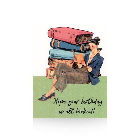 This birthday card features an oversized stack of books with an illustrated lady leaning up against the stack of books.   She is wearing black shoes, brown plaid pants, and a blue blazer and she is reading a book.  The background of the card is white and the base is green.  The message "Hope your birthday is all booked" is written in cursive font at the bottom of the card 