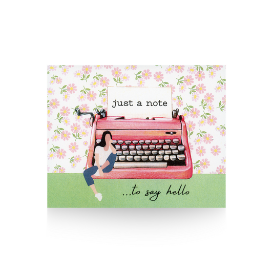This notecard features a pink flowered print background and green base.  On the base sits an illustration of a pink retro typewriter with a piece of paper in it that says "just a note" and sitting on the base of the typewriter is a modern woman with blue jeans and a white top.  At the bottom right hand corner of the notecard is written "...to say hello."
