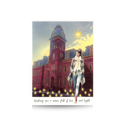 This holiday card features a wintry view of Woodburn Hall on the campus of West Virginia University.  A string of fairy lights is at the bottom of the photo.  A woman with blue jeans, a cream sweater and scarf, and tan overcoat is walking in front of the building and she has white lights around her as well. 