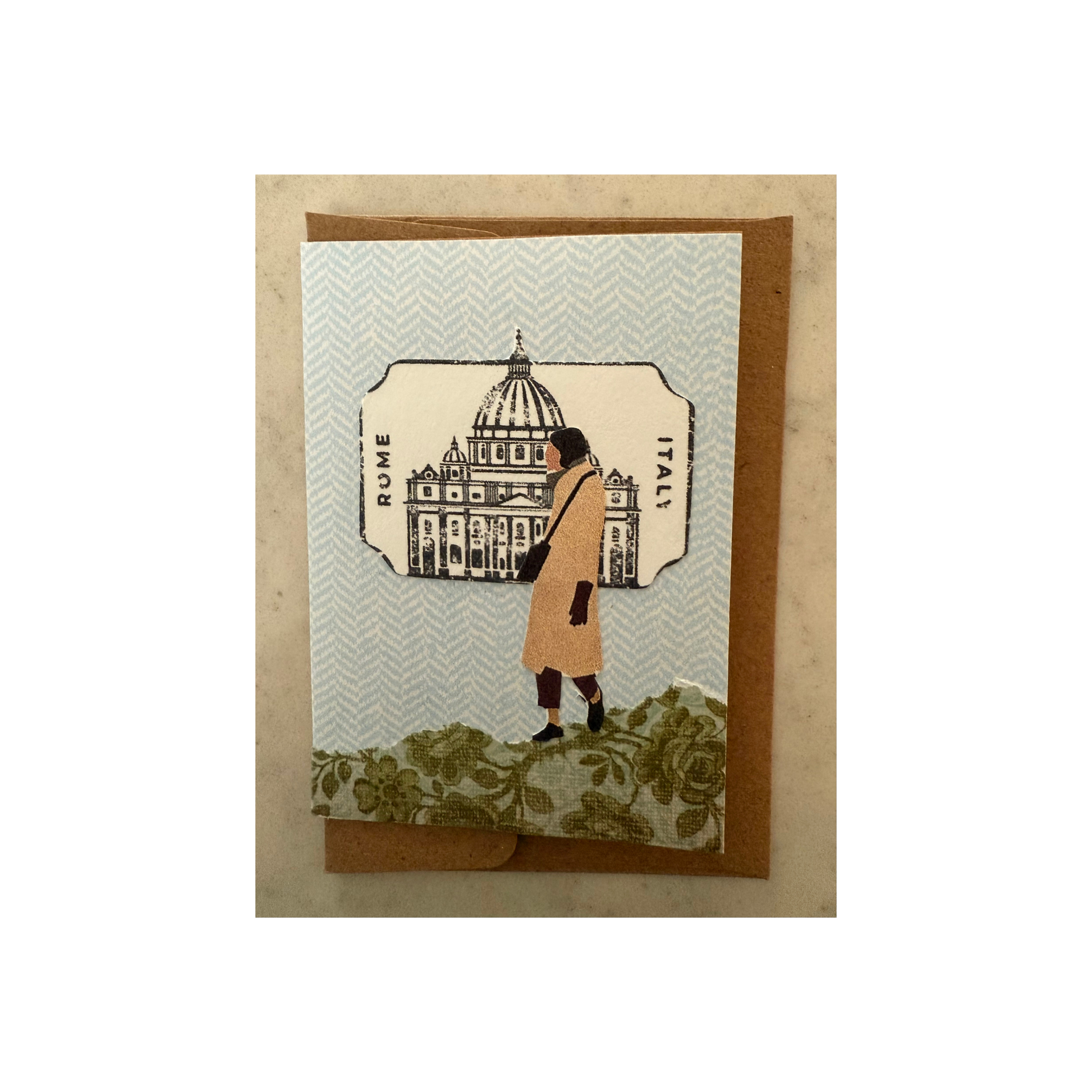 This mini collaged card is made of paper and features a lady in a camel-colored coat walking on a hill of green print paper with a blue print paper as background.  There is a cutout behind her of an Italian building