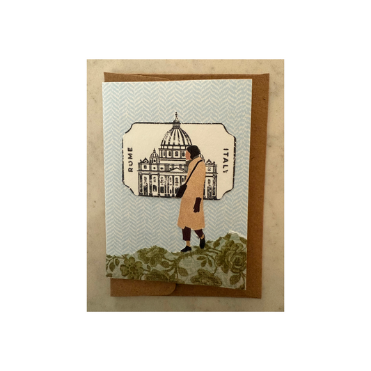 This mini collaged card is made of paper and features a lady in a camel-colored coat walking on a hill of green print paper with a blue print paper as background.  There is a cutout behind her of an Italian building